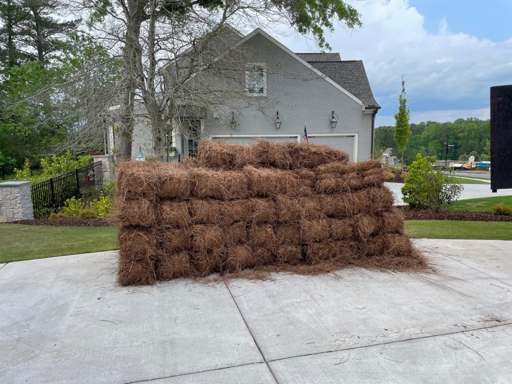 A wall of pine straw bales stacked on the driveway of a gray estate.
