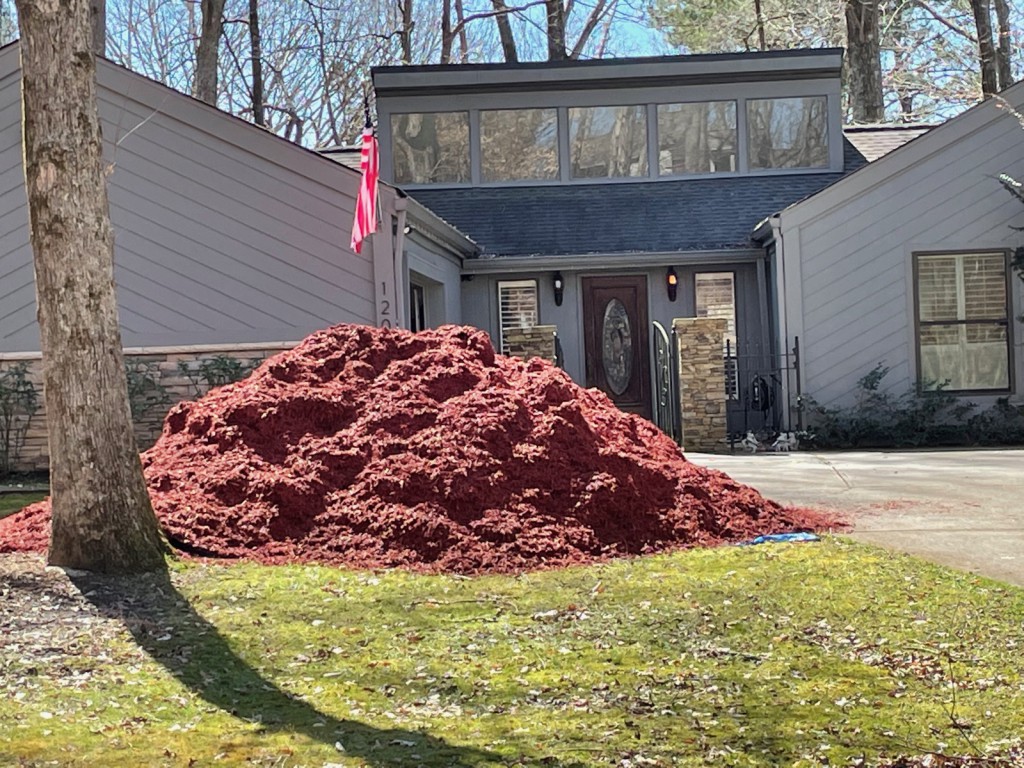 A tall red mulch pile delivered beside the driveway of a home flying an American flag.